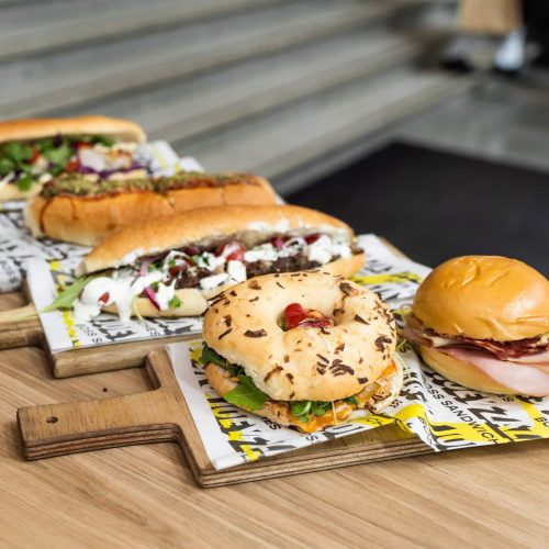 Joey Zaza's got sandwich rolls, bagels, and smooth burger buns for all 'ya sandwich catering needs. Featuring front to back: The Classic Conti in burger bun, Philly Cheesesteak in bagel, and sandwich rolls Big Bang Rendang and Mean Boss Parma.